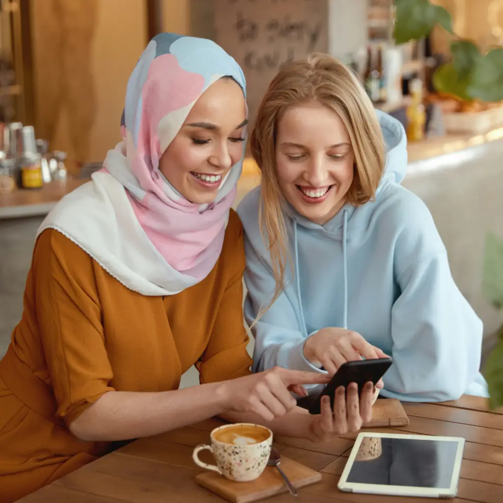 Two women friends in a café in Abu Dhabi, UAE, looking at a phone and connecting over parenting tips and support in an Eklektik Mama mum WhatsApp group